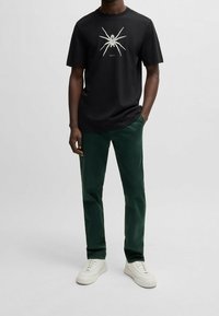 Czarny bawełniany t-shirt z białym nadrukiem pająka i logo, zestawiony z zielonymi sztruksowymi spodniami i białymi trampkami, stojący na jasnym tle.