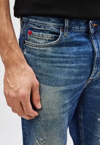 Falmede blå denimjeans med lett slitt finish, med fem lommer og et lite rødt logomerke på frontlommen.