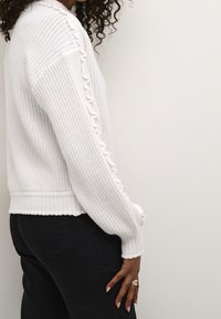 Witte gebreide trui met een textuurontwerp, met gerafelde accenten langs de mouw en geribde boorden en zoom. Gecombineerd met een zwarte broek.