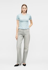 Γυναίκα με ίσια καστανέρυθρα μαλλιά, που φοράει μπλε κοντομάνικη μπλούζα με υφή, γκρι τζιν ψηλόμεσο και μαύρες γόβες με χαμηλό τακούνι.