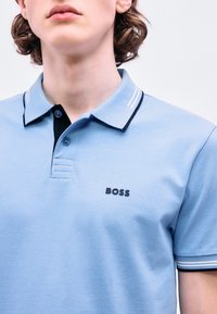 Νεαρός που φοράει ανοιχτό μπλε πόλο μπλούζα με σκούρες μπλε λεπτομέρειες και το λογότυπο "BOSS" στο στήθος, εμφανιζόμενος από τον λαιμό έως το μέσο του κορμού.