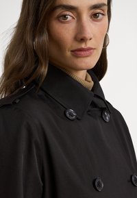 Czarny trencz o dwurzędowym kroju, z zaokrąglonymi czarnymi guzikami i wyprofilowanym kołnierzem, zestawiony z beżową bluzką z wysokim golfem.