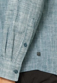 Close-up van een blauw-witte geblokte overhemdmouw met knopen, met gedetailleerde stoftextuur en een klein stoflabel nabij de zoom.