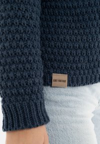 Marine gebreide trui met een gestructureerd steekpatroon en geribbelde manchetten. Inclusief een beige label met de tekst "Knit Factory" aan de onderkant.