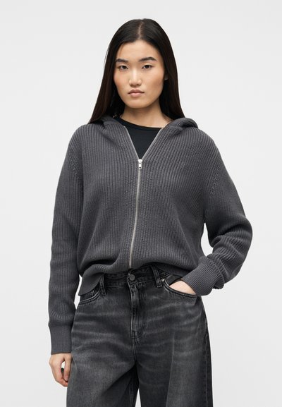 Jeune personne portant un sweat à capuche en tricot gris foncé avec fermeture éclair et un jean noir délavé, posant avec une main dans la poche sur un fond uni.