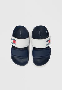 Sandalias tipo slide en azul marino con correas blancas ajustables que incluyen un logo rojo y azul. Plantilla texturizada con superficie lisa. Diseño ligero.