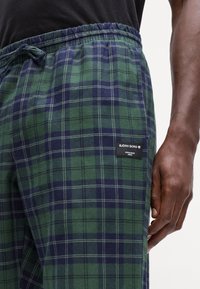 Close-up van een persoon die groene en marineblauwe geruite broek draagt met een Björn Borg-label op de dij, hand naast het been rustend.