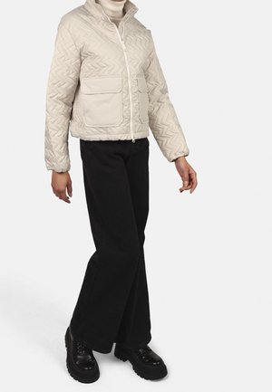 Lichtbeige gewatteerde jas met een hoge kraag, twee voorkantzakken en een ritssluiting, gecombineerd met zwarte wijde broek en zware zwarte laarzen.