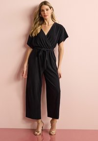 Svart plisserad jumpsuit med djup V-ringning, korta ärmar och ett knytbälte i midjan. Kombinerad med strappy guldfärgade högklackade sandaler.