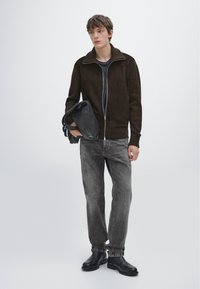 Brun zip-up jacka med ribbad textur, grå jeans och svarta läderstövlar. Modellen håller en svart läderväska med remmar.