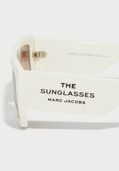 Ochelari de soare albi cu o ramă groasă, având text negru cuprinzând "THE SUNGLASSES" și "MARC JACOBS" pe brate. Lentile semi-opace.