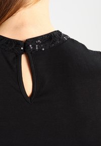 Blouse noire avec un col rond orné d'une garniture en dentelle décorée de sequins et une ouverture en forme de goutte à l'arrière. Texture de tissu lisse.