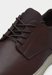 Chaussure en cuir marron avec des lacets ronds marron foncé, des coutures visibles et une semelle de couleur claire, présentée en gros plan sur un fond blanc.