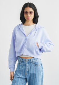 Κοντό φούτερ με φερμουάρ σε παστέλ λιλά, κατασκευασμένο από απαλό ύφασμα με μπροστινή τσέπη και διακριτικό λογότυπο. Συνδυάζεται με ανοιχτόχρωμα τζιν με ρίγες.