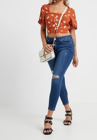 Hnědý oranžový crop top s květinovým vzorem a čtvercovým výstřihem, doplněný o roztrhané modré skinny džíny. Černé sandály na vysokém podpatku a bílá kabelka.