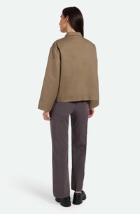 Chaqueta marrón con un corte amplio, que presenta un dobladillo recto y sin cierres, combinada con pantalones grises y zapatos negros. Vista trasera.