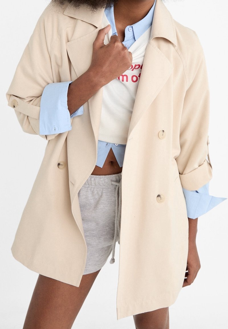 Trench-coat beige à double boutonnage avec manches retroussées, associé à une chemise bleu clair à col et à un short gris. Doté de boutons simples.