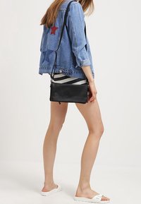 Czarna skórzana torba na ramię z długim paskiem, o gładkiej fakturze i minimalistycznym designie, zestawiona z denimową kurtką i casualowym obuwiem.