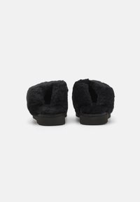 Chaussons noirs en peluche avec un dos ouvert et une texture douce, présentant une semelle plate et un intérieur rembourré pour plus de confort.