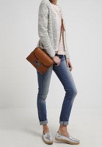 Sac à bandoulière en cuir marron avec ferrures argentées, cardigan gris texturé, chemise rayée, jean bleu aux revers retroussés, et chaussures plates métalliques.