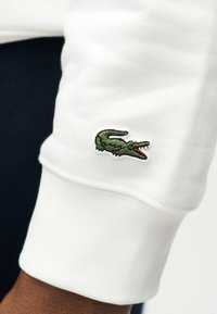 Manche en tissu blanc avec un logo de crocodile vert brodé près du poignet sur un bras à la peau foncée.