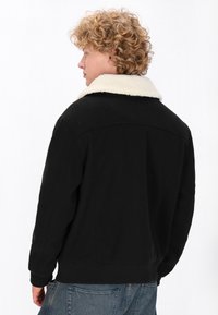 Sort jakke med en cremefarvet kunstpelskrave, der har et struktureret design og ribbede manchetter. Bagsiden har minimal pynt.