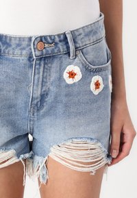 Shorts en denim bleu clair avec un ourlet effiloché et des fleurs blanches brodées avec des cœurs orange sur le devant, dotés d'un bouton en métal et d'une fermeture éclair.