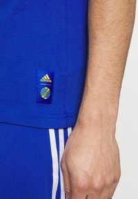 Modré sportovní tričko s krátkými rukávy, s logem Adidas a nášivkou s kulatým vzorem přišitou na spodní straně.