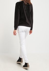 Sudadera de forro polar negra, jeans ajustados blancos y zapatillas negras con acentos en bronce. Texturas suaves y diseño casual visibles desde atrás.