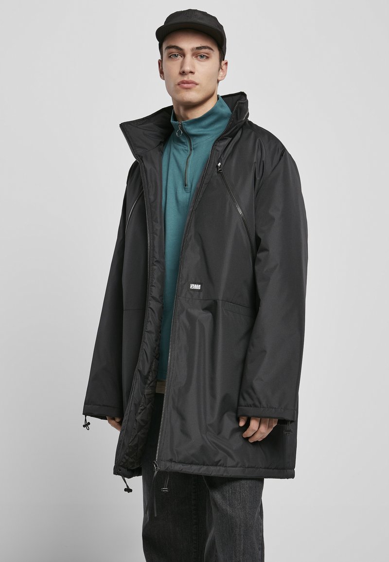 Svart vanntett lang frakk med høyt krage, glidelåser på sidene og justerbar fald. Brukt over en teal zip-up genser og mørke bukser.