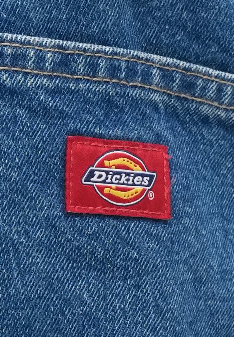 Red rectangular fabric tag with the logo "Dickies" in blue text, sewn onto blue denim fabric with visible stitching and texture.