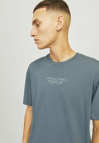 Katoenen T-shirt in gedempt blauw met korte mouwen, ronde halslijn en een centraal logo in witte en rode tekst. Glad textuur.