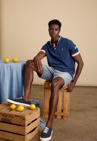 Marineblauwe polo met roze kraagdetails, denim shorts en marineblauwe sneakers. Citroengele accenten op een blauwe tafelkleed en houten kisten.