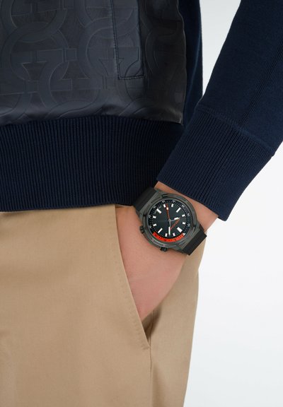 Black watch with orange accents and white indicators on a round dial. Worn on a wrist, paired with a navy sweater and beige trousers.