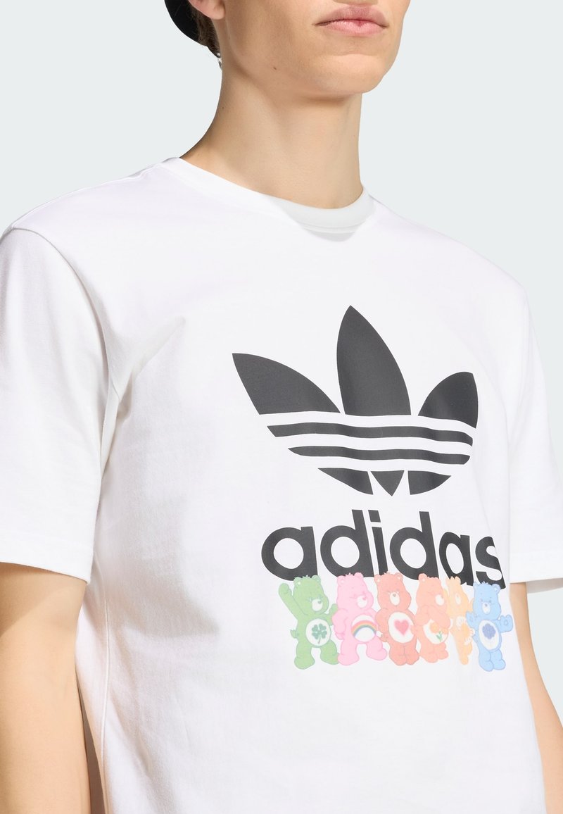 Person trägt weißes Adidas T-Shirt mit großem schwarzem Trefoil-Logo und sechs bunten Care Bears, die unter dem Markennamen gedruckt sind.