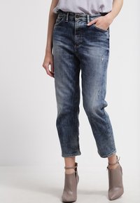 Kvinna som bär bleka blå, korta jeans med lätt slitningar och taupe ankelstövlar med sidodragkedjor och ankelremmar.