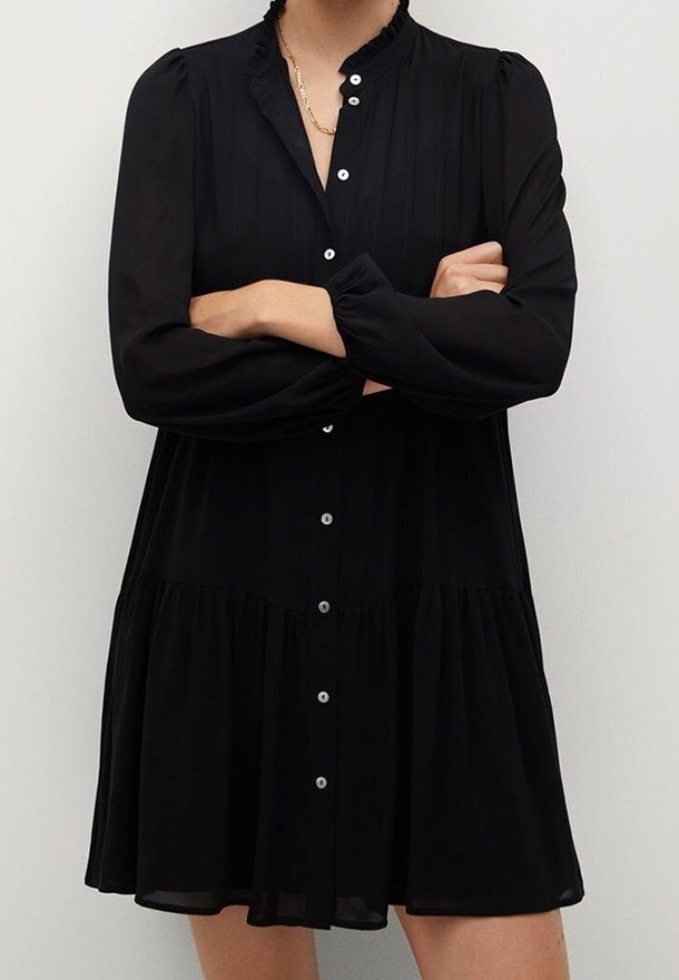 Robe noire à manches longues avec boutons sur le devant, jupe froncée et col à volants portée par une personne les bras croisés.