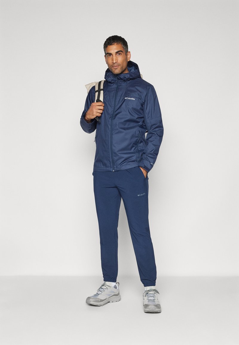 Marine wasserfeste Jacke mit verstellbarer Kapuze, Reißverschluss und Seitentaschen, kombiniert mit passenden Hosen und grauen Wanderschuhen.