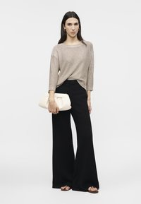Femme aux longs cheveux noirs portant un pull en tricot beige, un pantalon ample noir, tenant une grande pochette blanche, debout devant un fond blanc.