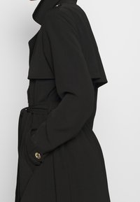 Trench-coat noir à double boutonnage, col large, grands revers et taille cintrée par une ceinture. Tissu lisse avec des boutons dorés en accents.
