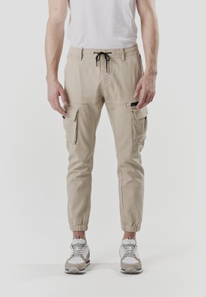 Pantalones cargo beige con cintura elástica, cordón y múltiples bolsillos. La tela es ligera con una textura suave, ajuste entallado.