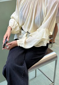 Osoba u bijeloj bluzici dugih rukava i crnoj suknji sjedi na metalnoj stolici, držeći pametni telefon s prstenjima na prstima.