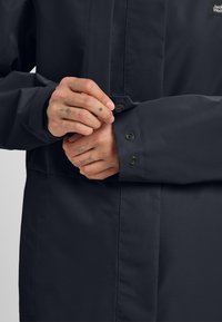 Dunkelfarbene, wasserdichte Jacke mit glatter Textur, die über Druckknopfverschlüsse an den Ärmelbündchen und ein minimalistisches Design verfügt.