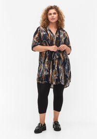Tunique fluide bleu marine avec un motif abstrait orange ; elle a des manches courtes, un col en V et des boutons. Portée sur des leggings noirs et des mocassins.