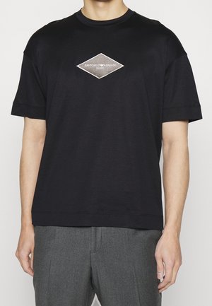 T-shirt en coton noir avec un col rond, des manches courtes et un logo en forme de diamant argenté au centre, avec des accents brodés.