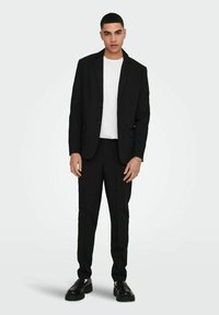 Giacca e pantaloni neri abbinati a una t-shirt bianca, con un design elegante e vestibilità su misura. Le scarpe nere completano il look.