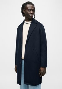 Manteau en laine bleu marine avec un col cranté, une fermeture à un bouton et des poches latérales, porté sur un pull léger et un jean.