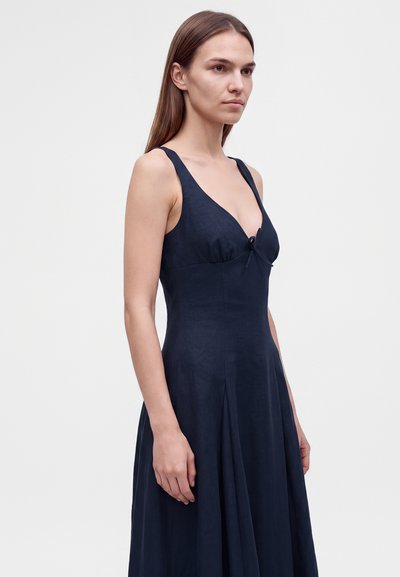 Γυναίκα με ίσια καστανά μαλλιά που φοράει αμάνικο φόρεμα σε σκούρο μπλε χρώμα με βαθύ ντεκολτέ σε σχήμα V, στέκεται μπροστά σε απλό ανοιχτό φόντο.