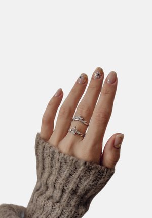 Dos anillos de plata con acentos de piedras preciosas se llevan en una mano que muestra un arte de uñas floral. La mano está parcialmente cubierta por un suéter gris texturizado.