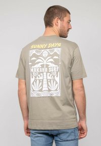 T-shirt vert olive avec un design graphique blanc comprenant un palmier, un soleil et le texte "JOURS ENSOLEILLÉS MAKAKA SURF" au dos. En matière coton.